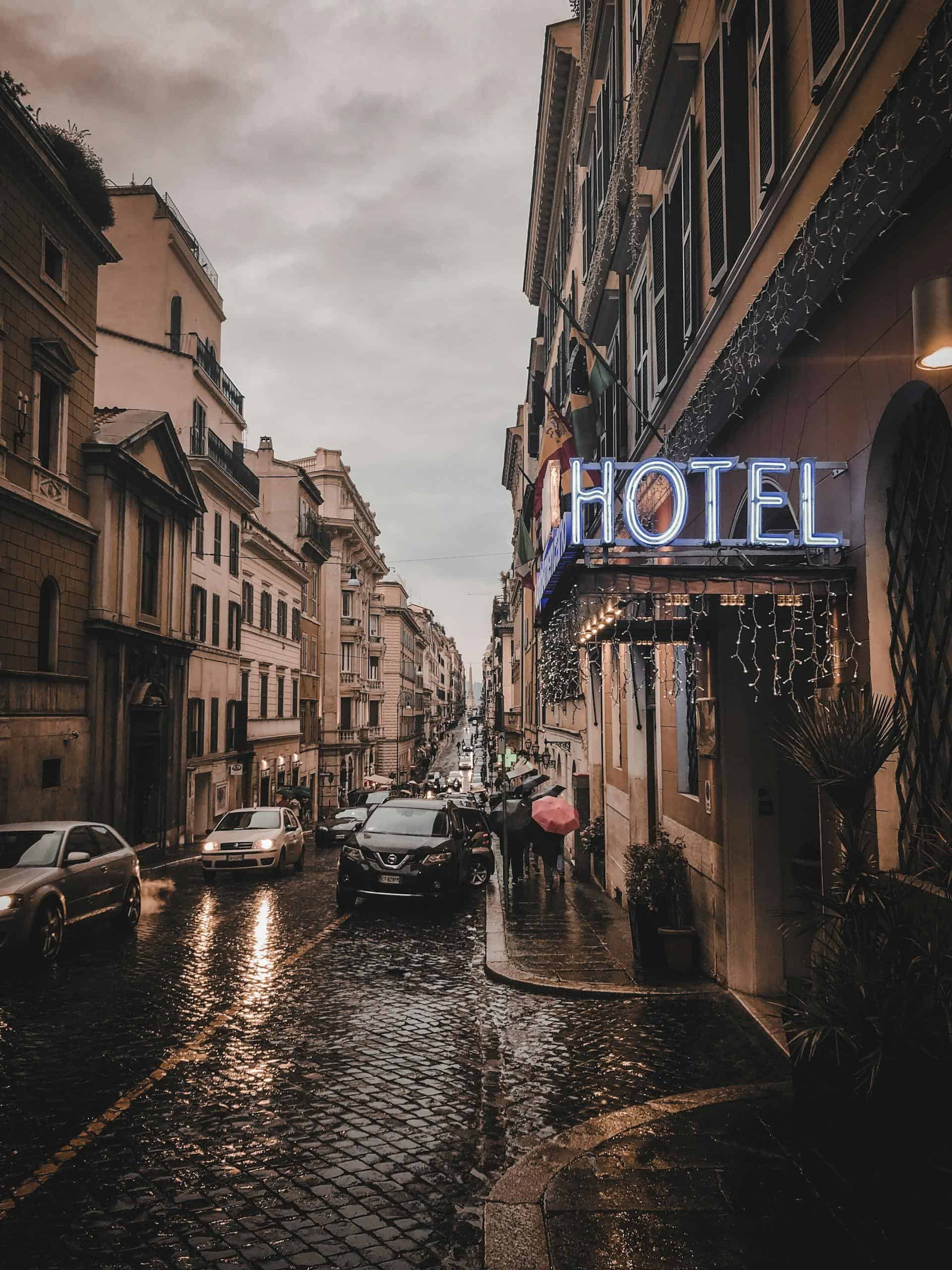 A rainy city street scene at dusk, featuring a glowing neon hotel sign, wet cobblestones, and bustling cars.