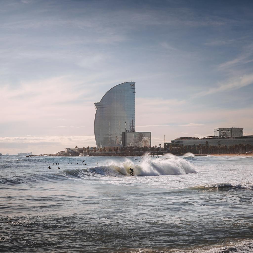 Surfer navigating waves with the backdrop of the iconic W Barcelona Hotel on a sunny day.
